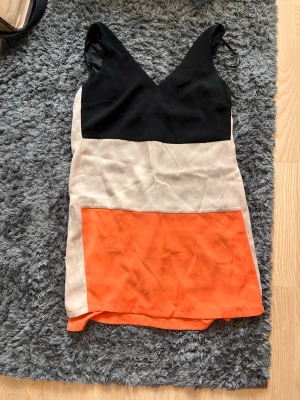 Blockfärgad ärmlös klänning - Snygg kortklänning med blockfärger i svart, beige och orange. Klänningen är ärmlös med v-ringad hals och har en rak passform. Tillverkad i ett lätt och mjukt material som ger en skön känsla. Perfekt för dig som gillar färg och enkel design.