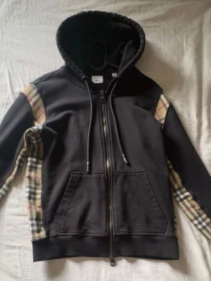 Svart Burberry hoodie - Säljer en svart hoodie från Burberry med dragkedja och klassiska beige-rutiga paneler längs ärmarna. Hoodien har huva och känguruficka framtill. Perfekt för dig som gillar streetwear med lyxig touch.