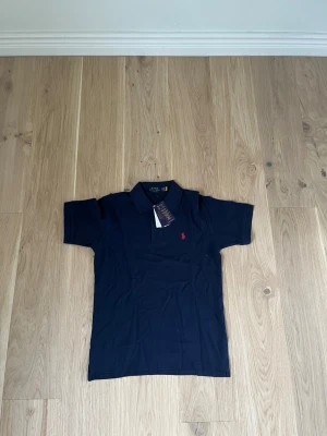Mörkblå pikétröja från Ralph Lauren - Klassisk mörkblå pikétröja från Ralph Lauren med korta ärmar och röd broderad logga på bröstet. Tröjan har krage och knappar vid halsen. Perfekt till en avslappnad men ändå stilren look. Har 2 st just nu i denna färg 