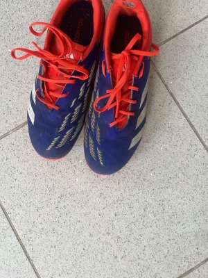 Blå och röda Adidas fotbollsskor Elite - Säljer ett par snygga fotbollsskor från Adidas i blått med röda detaljer och vita ränder. Skorna har klassisk låg profil, snörning och slitstark syntetisk ovandel. Perfekta för dig som vill sticka ut på planen!