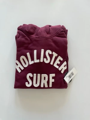 Helt ny, Vinröd hoodie från Hollister - HELT NY med prislapp, Snygg vinröd hoodie från Hollister med stor vit broderad text 'HOLLISTER SURF' på bröstet. Klassisk huva och mjukt material som känns skönt mot huden. 