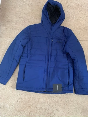 Blå dunjacka från Peak Performance - Snygg blå dunjacka från Peak Performance med huva och dragkedja framtill. Jackan har en bröstficka med dragkedja och diskret logga på bröstet samt ärmen. Perfekt för kalla vinterdagar tack vare det värmande dunmaterialet.