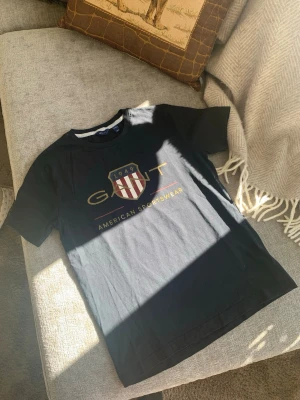 Mörkblå GANT t-shirt med tryck - Snygg mörkblå t-shirt från GANT med stort tryck framtill i gult och rött, samt ett sköldmärke med texten '1949' och 'American Sportswear'. Klassisk passform och rund halsringning. Perfekt för dig som gillar stilrena och sportiga plagg. Storlek 158-164 cm,  14-15 år. 