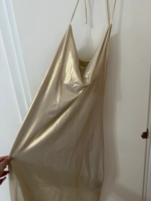 Champagnefärgad satin klänning - Säljer en glansig champagnefärgad klänning i satin med tunna axelband och draperad urringning. Klänningen har en slimmad passform och är perfekt för dig som vill sticka ut. Enkel men elegant design som ger en lyxig känsla.