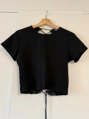 Svart topp med snörning och spets - Cool svart topp med korta ärmar och snörning i ryggen. Baksidan har dekorativ spets och öppet parti som ger en unik look. Perfekt för dig som vill sticka ut och gillar detaljer. Materialet känns som bomull och toppen är croppad.