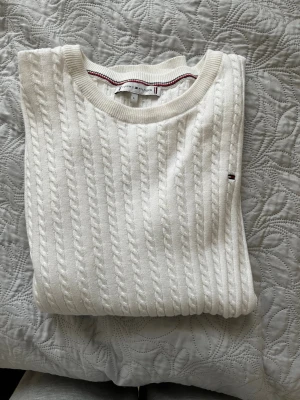 Vit kabelstickad tröja Tommy Hilfiger - Kabelstickad vit tröja från Tommy Hilfiger med rund halsringning och diskret logga på bröstet. Tröjan har ribbade muddar och är gjord i mjukt material som känns skönt mot huden. Perfekt för dig som gillar klassisk och stilrent. Den är i princip ny då den är använd endast 1-2 gånger. Därför mycket bra skick 