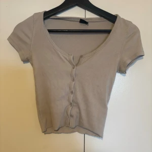 Beige ribbad croppad topp med knappar - Trendig beige croppad topp med korta ärmar och ribbad struktur. Toppen har en djup rundringning och knappar framtill som ger en snygg detalj. 