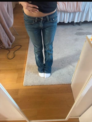 Blå bootcut jeans med låg midja - Säljer ett par klassiska blå bootcut jeans med låg midja och fem fickor. Jeansen har en snygg tvätt och subtila slitningar för en avslappnad look. Passar perfekt till en trendig vardagsstil och är tillverkade i mjukt denimtyg.