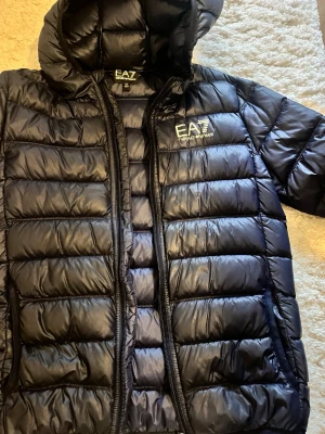 Svart pufferjacka från EA7 Emporio Armani - Snygg blå pufferjacka från EA7 Emporio Armani med huva och dragkedja. Jackan har ett stilrent quiltat mönster och logga på bröstet. Perfekt för kalla vinterdagar och ger en trendig look. Tillverkad i glansigt material som känns lätt men ändå värmande.