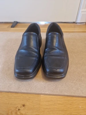 Svarta loafers i skinn ECCO - Säljer ett par klassiska svarta loafers i skinn med fyrkantig tå och diskret söm längs ovandelen. Skorna har en robust gummisula med tydligt mönster för bra grepp. Perfekta för dig som gillar stilrena och tidlösa skor.