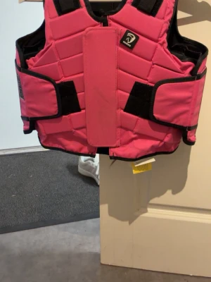Rosa säkerhetsväst från Riders Company - Säljer en rosa säkerhetsväst från Riders Company med svart kant och justerbara kardborreband. Västen har vadderad design och är perfekt för ridning. Märket syns tydligt framtill och västen har en sportig look den är max använd 6-7 gånger men inte mer den är sparsamt använd har köpt den för 1325kr men säljer den för 679kr den är i storlek XXS-XS i barn storlek 