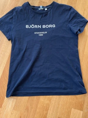 Marinblå Björn Borg t-shirt - Snygg marinblå t-shirt från Björn Borg med vit text på bröstet där det står 'BJÖRN BORG STOCKHOLM 1956'. Klassisk rund halsringning och korta ärmar. Tillverkad i mjuk bomull som känns skön mot huden.