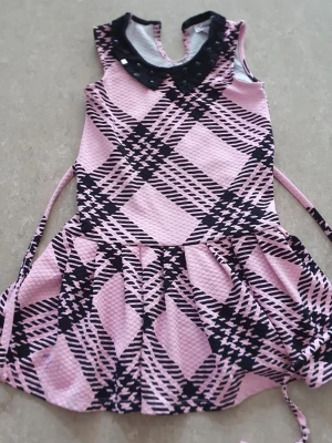 Klänning - Söt klänning i rosa och svart med rutigt mönster och dekorativ svart krage med små pärlor. Klänningen har ärmlös design och knytband i midjan för extra passform. Perfekt för dig som gillar en lekfull och trendig stil. Fläckar av bläck.