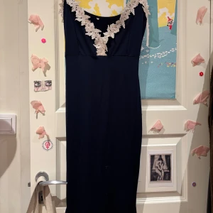 Marinblå långklänning med spets - Elegant marinblå långklänning med v-ringning och dekorativ vit spets längs urringningen. Klänningen har smala axelband och en figurnära passform som ger en stilren siluett. Perfekt för speciella tillfällen där du vill känna dig extra fin.