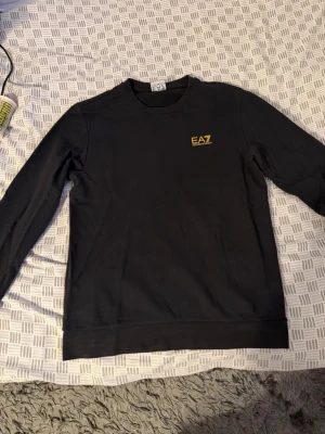 Svart sweatshirt från EA7 Emporio Armani - Säljer en svart sweatshirt från EA7 Emporio Armani med gul logga på bröstet. Tröjan har rund halsringning och långa ärmar. OBS! Finns små vita knappt synliga fläckar vid armhålorna samt lite vid kanten längst ner.