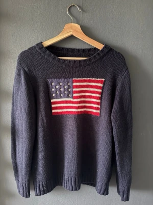 Mörkblå stickad tröja med USA-flagga - Stickad tröja i mörkgrå färg med amerikansk flagga framtill. Väldigt skön och mjuk att ha på sig, perfekt till vardags. Klassisk modell med rund hals och ribbade muddar vid ärmar och nederkant. Använd men i fint skick, inga större defekter vad jag kan se. Passar bra till jeans eller leggings för en avslappnad stil. Skirv om du har frågor!💞