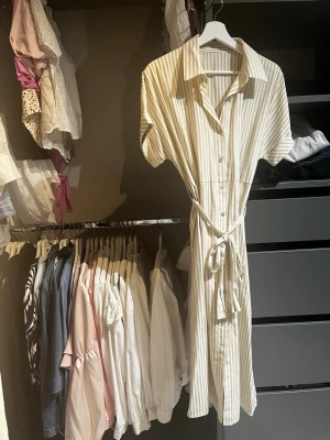 Randig skjortklänning med knytband - Snygg kort skjortklänning med beige och vita ränder. Klänningen har korta ärmar, knappar framtill och ett knytband i midjan för en smickrande passform. Perfekt för en avslappnad och trendig look.