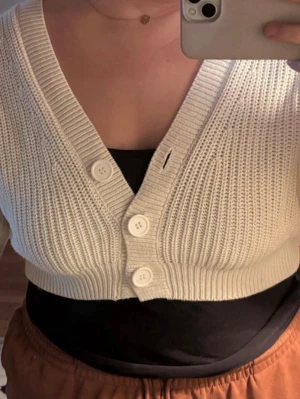 Vit cropped kofta från Divided - Säljer en vit, ribbstickad cropped kofta från Divided (HM) med tre stora knappar framtill. Koftan har djup V-ringning och breda muddar. Perfekt att slänga över ett linne eller t-shirt för en chill look.