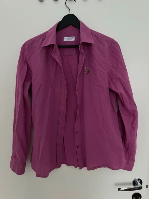 Rosa skjorta från US POLO ASSN - Stilren rosa skjorta från US Polo Assn med klassisk krage och lång ärm. Skjortan har knappar framtill och en liten broderad logga på bröstet. Perfekt för dig som vill ha en färgklick i garderoben.