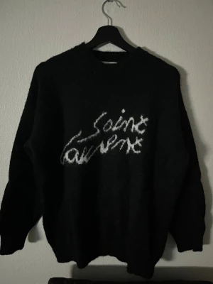 Svart Saint Laurent stickad tröja - Säljer en svart stickad tröja från Saint Laurent med vit logotyp i grafisk stil på bröstet. Tröjan har rund halsringning, långa ärmar och ribbade muddar. Perfekt för dig som vill ha en trendig och exklusiv look.