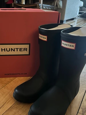 Svarta gummistövlar från Hunter - Säljer ett par klassiska svarta gummistövlar från Hunter med logga framtill och knäppe på sidan. Stövlarna har mellan höjd. 