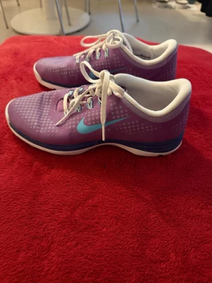 Lila Nike sneakers med turkos swoosh - Snygga lila Nike sneakers med turkos swoosh och vita detaljer. Skorna har vita snören, vit insida och en blå rand längs sulan. Ovandelen är i mesh-liknande material med små vita prickar för extra stil. Perfekta för dig som vill ha färg på fötterna!