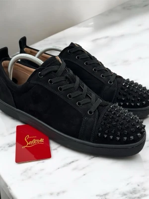 Svarta sneakers från Christian Louboutin - Snygga svarta sneakers i mocka från Christian Louboutin med svarta nitar på tån och klassisk röd sula. Skorna har svarta skosnören och en stilren design som sticker ut med sina detaljer. Perfekta för dig som vill ha något unikt, lyxigt och trendigt. Storleken är 43, den var för liten därför säljer de. 