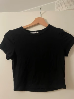 Svart croppad t-shirt från Zara - En enkel och stilren svart croppad t-shirt från Zara. T-shirten har rund halsringning och korta ärmar. Tillverkad i mjuk bomull som känns skön mot huden. Perfekt till jeans eller kjol för en clean look.