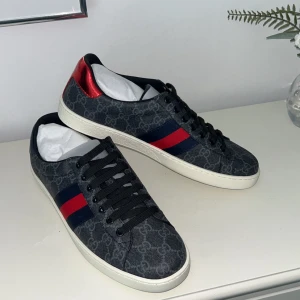 Gucci sneakers med GG-mönster - Snygga Gucci sneakers i mörkgrått med klassiskt GG-mönster, röda och marinblå ränder på sidorna och metallicröd samt metallicblå detalj på hälen. Skorna har svarta snören och vit platt sula. Perfekta för dig som vill sticka ut med lyxig streetstyle. Hör av er om ni har funderingar. Kom också med prisförslag!