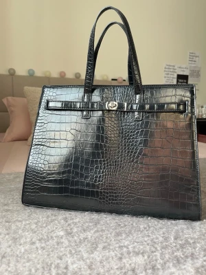 Svart handväska i krokopräglat läder - Stilren svart handväska i krokopräglat läder med två topphandtag och elegant guldfärgat spänne framtill. Väskan har en fyrkantig form och en lyxig finish som ger en exklusiv känsla. Perfekt accessoar för dig som gillar trendiga detaljer.