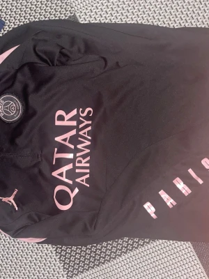 Svart PSG träningströja Jordan - Snygg svart träningströja från PSG i samarbete med Jordan. Tröjan har rosa detaljer, PSG-logga på bröstet och Qatar Airways-tryck framtill. Tillverkad i ett lätt och funktionellt material som passar perfekt till träning eller match.