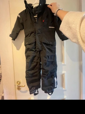 Svart täckoverall från Didriksons - Säljer en svart täckoverall från Didriksons med huva och reflexdetaljer. Overallen har dragkedja framtill, fickor med lock och resår vid ärm- och benslut. Perfekt för kalla dagar och snölek. Materialet är slitstarkt och vattenavvisande.