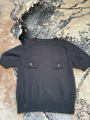 Svart stickad tröja med fejk fickor - Snygg svart stickad tröja med korta ärmar och två dekorativa fickor framtill med knappdetaljer. Tröjan har ribbade muddar och ett diskret hålmönster längs framsidan. Perfekt för lager på lager och en chill look. Tyvärr har den ingen varumärkesettikett därför vet jag inte vart den är ifrån. Anledningen till att jag säljer den är på grund av att den inte är min stil längre. 