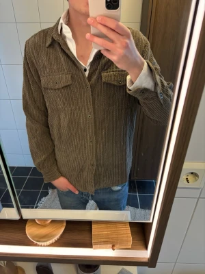 Brun manchester overshirt från True Grit - Säljer en brun overshirt i manchester från True Guy. Skjortan har långa ärmar, två bröstfickor med knappar och klassisk krage. Perfekt att bära öppen över en t-shirt eller skjorta för en avslappnad look.