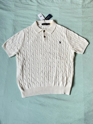Vit stickad polotopp från Polo Ralph Lauren - Vit stickad polotopp från Polo Ralph Lauren med korta ärmar, kabelstickat mönster och två knappar vid kragen. Klassisk pikékrage och broderad logga på bröstet. Perfekt för en stilren look.