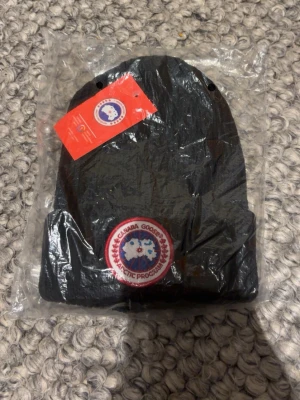 Svart mössa från Canada Goose - Svart ribbstickad mössa från Canada Goose med klassisk rund logotyp framtill. Mössan har uppvikt kant och är tillverkad i ett mjukt material som passar perfekt för kalla dagar. Snygg och enkel design som funkar till det mesta.