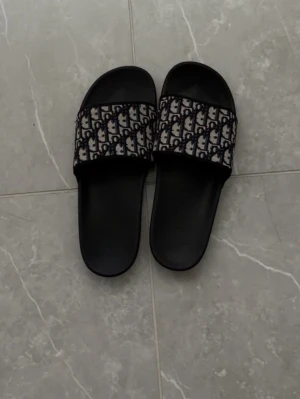 Svarta Dior tofflor med monogram - Säljer ett par svarta tofflor från Dior med klassiskt monogrammönster i vitt och blått på remmen. Tofflorna har en bred rem och en bekväm sula i gummi, perfekta för chill dagar hemma eller på stan.