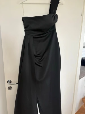 Svart långklänning med oneshoulder - Elegant svart långklänning med oneshoulder-design och draperad överdel. Klänningen har en stilren och modern look med slits framtill och är tillverkad i ett glansigt material som ger en lyxig känsla.