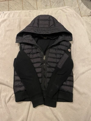 Moncler Cardigan  - Säljer en svart Cardigan från Moncler med stickade ärmar och huva. Jackan har quiltad front, dragkedja och Moncler-logga på ärmen. Perfekt för kalla dagar och riktigt snygg att matcha med jeans eller joggers. Priset är ej hugget i sten så skicka ett prisförslag så kan vi förhandla‼️‼️Nypris 7599kr‼️