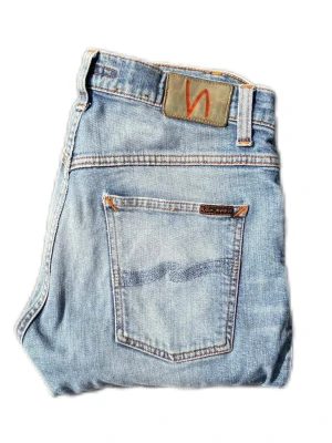 Ljusblå jeans från Nudie Jeans - Säljer ett par ljusblå jeans från Nudie Jeans med klassisk femficksdesign och slitning på höger ben. Jeansen har en slim fit och är gjorda i bomull med snygga detaljer som kontrastsömmar och Nudie-logga på bakfickan. Lagade i skrevet 