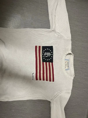 Vit stickad tröja från Bondelid - Vit stickad tröja från Bondelid med amerikansk flagga i rött, vitt och blått på bröstet samt broderad Bondelid-logga under flaggan. Tröjan har rund halsringning och långa ärmar. Perfekt för dig som gillar klassisk och avslappnad stil. SITTER SOM S