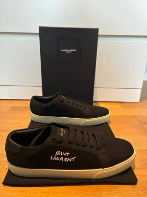 Saint Laurent sneakers - Svarta sneakers från Saint Laurent med vit broderad logga på sidan och vit sula. Skorna har klassisk låg profil, svarta skosnören och är tillverkade i canvas med detaljer i läder. Perfekta för en stilren och trendig look.