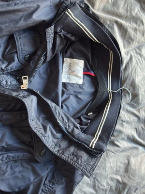 Mörkblå vindjacka från Moncler - Snygg mörkblå vindjacka från Moncler med dragkedja och ribbad krage med vita ränder. Jackan är tillverkad i lätt och slitstarkt material, perfekt för blåsiga dagar. Klassisk design med långa ärmar och diskreta detaljer. Kommer ej kunnas pruta så strunta skriv om ni ej är intresserad på riktigt 
