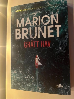 Grått hav av Marion Brunet - En spännande roman av Marion Brunet med omslag i mörkblått och grönt, föreställande ett stormigt hav och en ensam segelbåt. Boken är på svenska och utgiven av Gilla Böcker. Perfekt för dig som gillar dramatiska berättelser och havstema.