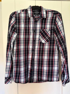 Rutig långärmad skjorta Bondelid - Snygg rutig skjorta från Bondelid i blått, vitt, grått och rött. Skjortan har klassisk krage, bröstficka med broderad logga och knäppning framtill. Perfekt för en avslappnad men stilren look.