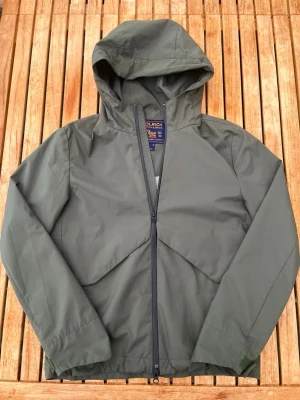 Grön vindjacka från Woolrich - Snygg grön vindjacka från Woolrich med huva och dragkedja framtill. Jackan har en minimalistisk design och är tillverkad i ett lätt, vindtätt material som passar perfekt för blåsiga dagar. Märkeslapp på insidan och justerbar nederkant för extra komfort.
