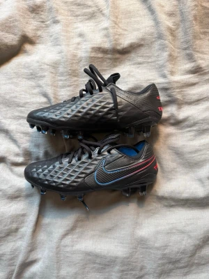 Nike Tiempo svarta fotbollsskor - Säljer ett par svarta Nike Tiempo fotbollsskor med blå och röda detaljer. Skorna är aldrig använda utan endast testade. 
