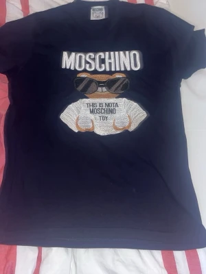 2 Svarta Moschino t-shirts med björn - Två svarta t-shirts från Moschino med broderad björn och texten 'This is not a Moschino toy' på bröstet. En har björnen med solglasögon och vit text, den andra med orange text och klassisk björn. Perfekt statement-plagg för dig som gillar streetstyle.