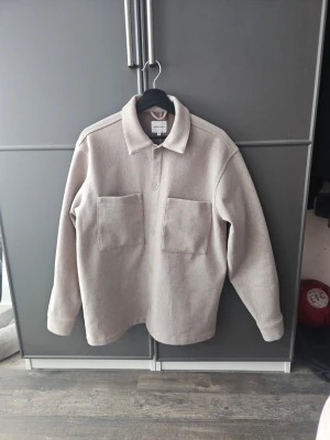 Beige overshirt från Jack and Jones - Stilren beige overshirt från Jack and Jones med två stora bröstfickor och klassisk krage. Overshirten har rak passform och stängs med dolda knappar framtill. Tillverkad i ett mjukt, tjockare tyg som passar perfekt för lager på lager.