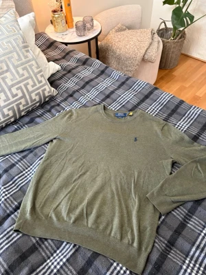 Polo Ralph Lauren tröja - Snygg färggrön tröja från Polo Ralph Lauren med rund halsringning och diskret broderad logga på bröstet. Tröjan är tunnstickad och har långa ärmar samt ribbade muddar vid ärmslut och nederkant. Perfekt för en stilren look. Liten defekt på taggen se på sista bild.